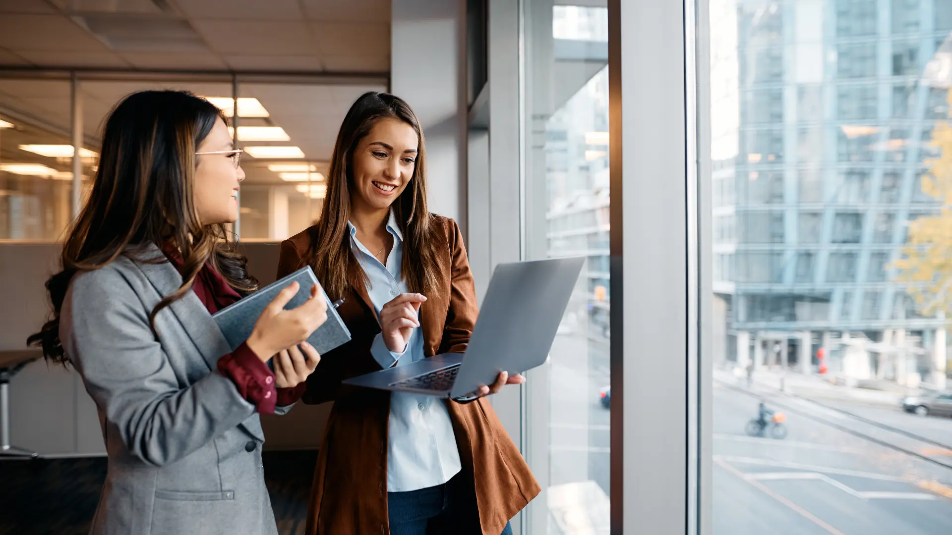 Two businesswomen reviewing a laptop by an office window
